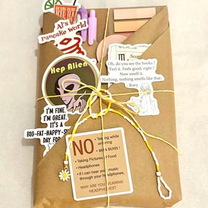 May include: A brown paper-wrapped package decorated with various stickers including a Long Island Iced Tea, Al's Pancake World, a green alien, and text quotes. The package is tied with yellow and white string featuring a small white daisy charm.
