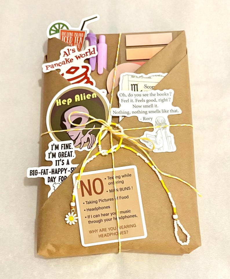 May include: A brown paper-wrapped package decorated with various stickers including a Long Island Iced Tea, Al's Pancake World, a green alien, and text quotes. The package is tied with yellow and white string featuring a small white daisy charm.