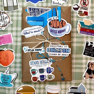 May include: A brown paper-wrapped package tied with twine, featuring a blue pen and various stickers including a Stars Hollow logo, a Chilton class of 2003 sign, a jumbo coffee mug, a burger, a house, a dog, and text-based quotes on a green grid.