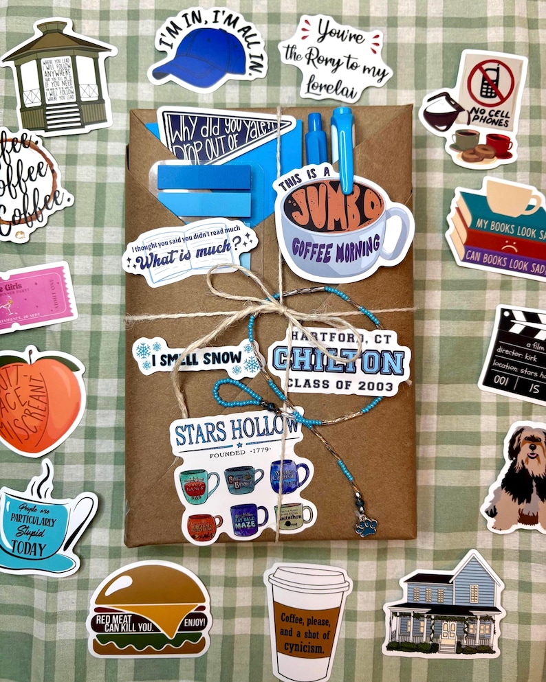 May include: A brown paper-wrapped package tied with twine, featuring a blue pen and various stickers including a Stars Hollow logo, a Chilton class of 2003 sign, a jumbo coffee mug, a burger, a house, a dog, and text-based quotes on a green grid.