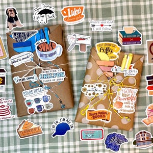 May include: Two brown paper-wrapped packages tied with string, one with blue and one with yellow pens tucked inside. The packages are surrounded by numerous colorful stickers featuring various illustrations and text related to the show Gilmore Girls.