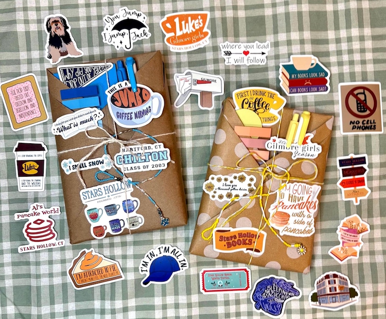May include: Two brown paper-wrapped packages tied with string, one with blue and one with yellow pens tucked inside. The packages are surrounded by numerous colorful stickers featuring various illustrations and text related to the show Gilmore Girls.