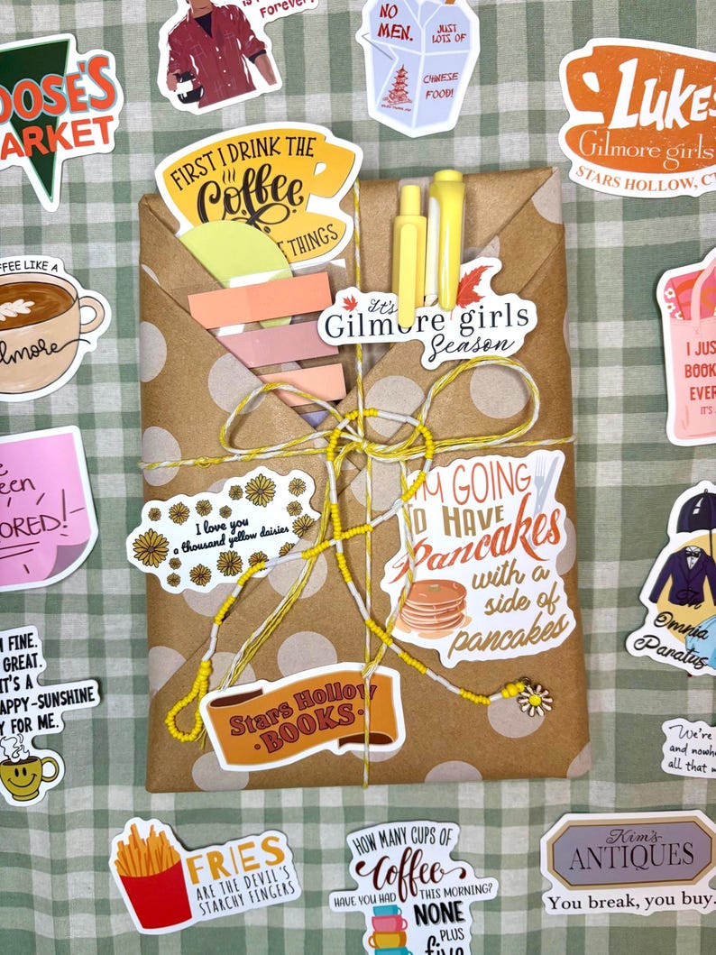 May include: A rectangular brown paper package with white polka dots, tied with yellow and white string. It features two yellow pens, a stack of pastel sticky notes, and stickers with text including First I Drink The Coffee, Gilmore Girls Season, I Love You A Thousand Yellow Daisies, I'm Going To Have Pancakes, and Stars Hollow Books.