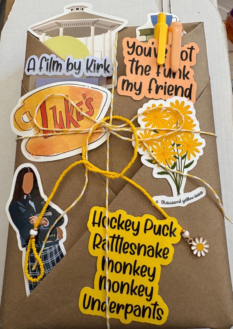 May include: A brown paper package decorated with various stickers including a gazebo, a coffee mug labeled Luke's, a bouquet of yellow daisies, and text reading A film by Kirk, You've got the funk my friend, and Hockey Puck Rattlesnake Monkey Monkey Underpants.