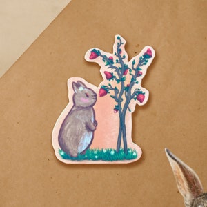May include: A gray rabbit with a white flower on its head is sniffing a pink strawberry on a green plant with multiple pink strawberries. The background is a soft pink.