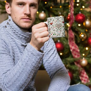 Puede incluir: Un hombre sostiene una taza de cer&aacute;mica blanca con un dise&ntilde;o navide&ntilde;o. La taza presenta un patr&oacute;n de corazones, adornos y copos de nieve rojos, verdes y blancos. El hombre lleva un su&eacute;ter gris y pantalones vaqueros azules. Est&aacute; sentado frente a un &aacute;rbol de Navidad decorado con adornos rojos y luces.