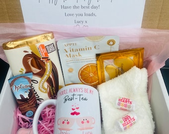 May include: A pink and white gift box filled with various items, including a mug with the text "You'll Always Be My Best-Tea" and two cartoon teacups, a chocolate bar, a face mask, a white towel, and pink heart-shaped candles. The box also includes two small candy rolls with the text "Love Hearts".
