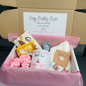 May include: A pink gift box filled with birthday gifts for a friend. The box contains a mug that says "You'll always be my best-tea", a white towel with a brown tag that says "Pocket hug for my bestie", a pink hair scrunchie, a chocolate bar, a bag of candy, and a card that says "Happy Birthday Bestie! Have the best day! Love you loads, Lucy x".