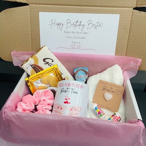 May include: A pink gift box filled with a variety of items, including a mug that says "You'll Always Be My Best-Tea", a chocolate bar, a white towel, a pink scrunchie, and a small card that says "Pocket Hug for My Bestie".