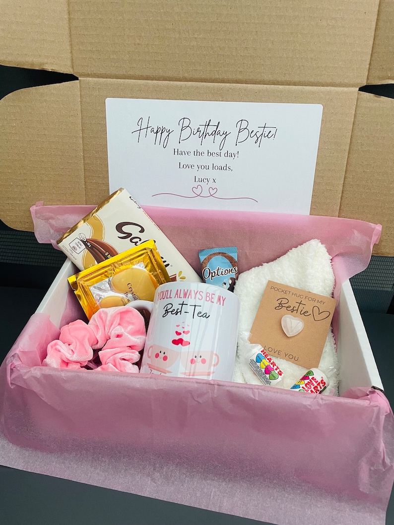 May include: A pink gift box filled with a variety of items, including a mug that says "You'll Always Be My Best-Tea", a chocolate bar, a white towel, a pink scrunchie, and a small card that says "Pocket Hug for My Bestie".