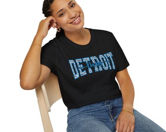 Könnte beinhalten: Schwarzes T-Shirt mit dem Wort "DETROIT" in blau-weißer Leopardenmuster-Schrift. Das Wort "Lions" ist kursiv in der Mitte des Wortes "DETROIT" geschrieben. Das Model trägt eine blaue Jeans.