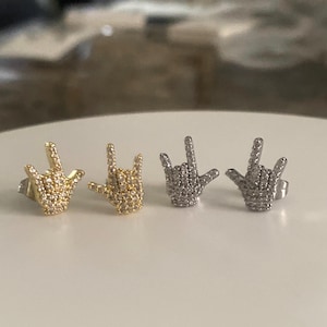 May include: Two pairs of stud earrings shaped like a hand making the I love you sign in American Sign Language. One pair is gold-toned and the other is silver-toned, both featuring small clear pavé stones covering the entire surface of the hand.