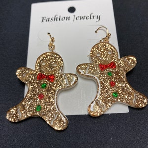 May include: Gold glitter gingerbread man earrings with red and green accents. The earrings have a gold hook and are on a white card that says "Fashion Jewelry". The earrings are a festive holiday accessory.