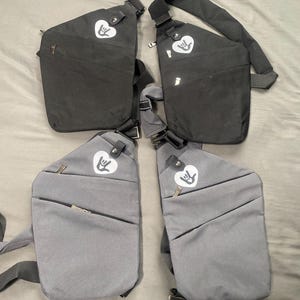 Könnte beinhalten: Vier Sling-Bags in Schwarz und Grau. Jede Tasche hat ein herzförmiges Logo mit einer Handgeste. Die Taschen haben eine Reißverschluss-Vordertasche und einen verstellbaren Riemen. Die Taschen sind aus strapazierfähigem Material.