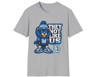 Könnte beinhalten: Graues T-Shirt mit einem Cartoon-Löwen, der eine blaue Mütze, Sonnenbrille und Turnschuhe trägt. Der Löwe hat blaues Fell und der Text "THEY NOT LIKE US" ist in Weiß und Blau dargestellt. Das Shirt hat auch den Text "LIONS 313".