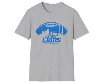 Könnte beinhalten: Graues T-Shirt mit blauem Football-Grafikdesign. Das Design zeigt das Logo der Detroit Lions und das Wort "FOOTBALL" darunter, mit einer Stadtsilhouette innerhalb der Footballform.