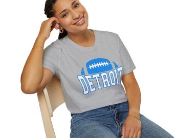 Könnte beinhalten: Hellgraues T-Shirt mit blauem Football-Motiv und dem Wort "DETROIT" in weißen Blockbuchstaben. Das Shirt besteht aus weichem Material und ist für den Freizeitgebrauch geeignet.