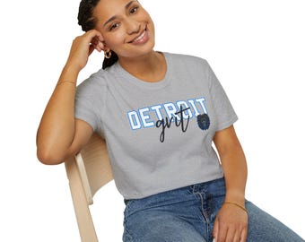Könnte beinhalten: Hellgraues T-Shirt mit den Worten "DETROIT grit" in blauer und schwarzer Schrift. Eine blaue Löwen-Grafik befindet sich rechts vom Wort "grit". Das T-Shirt wird von einer Person getragen.