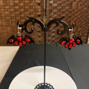 May include: A pair of black bat-shaped earrings with red rhinestone accents. The earrings are displayed on a black earring stand.