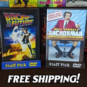 May include: Two DVD movie cases, "Back to the Future" and "Anchorman: The Legend of Ron Burgundy", displayed on stands labeled "Staff Pick DVD". The "Back to the Future" case features a man and a car. The "Anchorman" case features Will Ferrell.