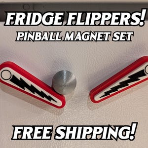 May include: Pinball-themed refrigerator magnet set. The set includes two red and white flipper magnets with black lightning bolt designs and a silver ball magnet. Text on the image reads "FRIDGE FLIPPERS! PINBALL MAGNET SET".