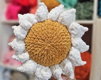 May include: A handmade, plush sunflower pillow with a golden-yellow center and white petals. The soft, textured pillow is designed to resemble a daisy, with a round center and petal-shaped edges. The pillow is held in a person's hand.