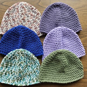 May include: A collection of six handmade knit beanies in various colors. The beanies include shades of blue, purple, green, and a multi-colored mix of brown, pink, and white. The hats are arranged on a wooden surface.