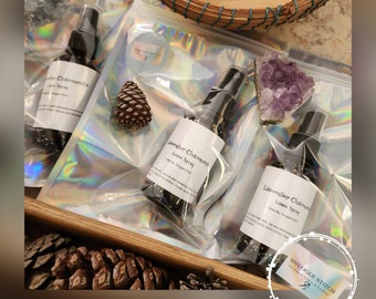 Peut inclure: Trois flacons pulvérisateurs noirs étiquetés "Lavender Chamomile Linen Spray" dans des sacs en plastique transparent. Les sacs sont sur une surface en bois avec des pommes de pin, des cristaux et un panier tressé. La marque "Snuggle Stitch" est visible.