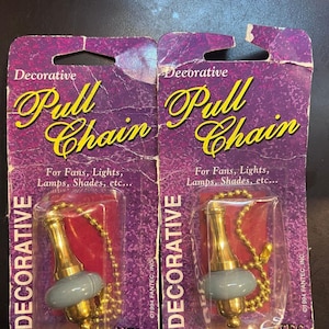 May include: Two packages of decorative pull chains for fans and lights. Each package features a gold-toned metal and grey ceramic pull handle attached to a gold ball chain, encased in clear plastic on a purple cardboard backing with yellow text.