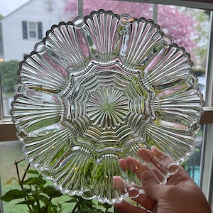 May include: Clear glass serving dish with a scalloped edge and a radial design. The dish has multiple compartments and a central, starburst-like pattern. The glass is transparent, allowing light to pass through, and is held by a person's hand.