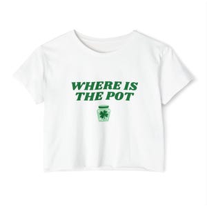 May include: White t-shirt with the text "WHERE IS THE POT" in green, above a green jar with a four-leaf clover. The shirt has a crew neck and short sleeves. The design is centered on the front of the shirt.