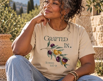 May include: A beige short-sleeved t-shirt featuring a graphic of an olive branch with green leaves and purple and gold olives. The text reads "GRAFTED IN." in dark green letters, with "ROMANS 11:17-18" printed in smaller black text below the branch.