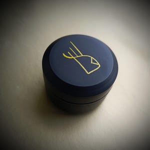 K&ouml;nnte beinhalten: Ein kleiner, runder, dunkelblauer Beh&auml;lter mit einem goldenen Design auf dem Deckel. Das Design ist ein stilisiertes Auge mit drei Linien, die daraus herausragen.