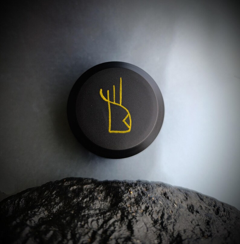 K&ouml;nnte beinhalten: Eine schwarze runde Scheibe mit einem goldenen Symbol darauf. Das Symbol ist eine stilisierte Hand mit drei Fingern und einer Diamantenform an der Basis.