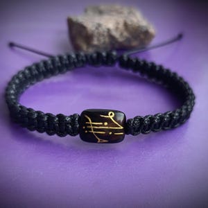 Puede incluir: Pulsera de macramé negra con una cuenta cuadrada marrón oscuro con símbolos dorados. La pulsera está sobre un fondo púrpura con una piedra. La pulsera es ajustable.