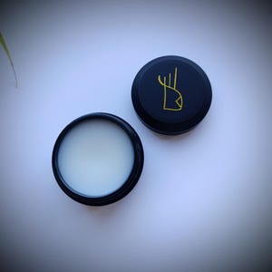 K&ouml;nnte beinhalten: Ein runder schwarzer Beh&auml;lter mit einer wei&szlig;en Creme darin. Der Deckel des Beh&auml;lters ist schwarz mit einem goldenen Design einer stilisierten Hand.