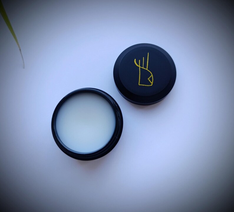 K&ouml;nnte beinhalten: Ein runder schwarzer Beh&auml;lter mit einer wei&szlig;en Creme darin. Der Deckel des Beh&auml;lters ist schwarz mit einem goldenen Design einer stilisierten Hand.