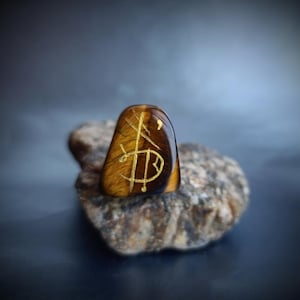 Puede incluir: Una piedra pulida de ojo de tigre con símbolos dorados grabados. La piedra, de forma ovalada, descansa sobre una roca gris rugosa. El fondo es un degradado borroso de azul oscuro y gris.