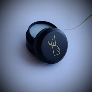 K&ouml;nnte beinhalten: Ein runder schwarzer Beh&auml;lter mit einem goldenen Design auf dem Deckel. Der Beh&auml;lter ist ge&ouml;ffnet und zeigt eine wei&szlig;e Creme im Inneren.