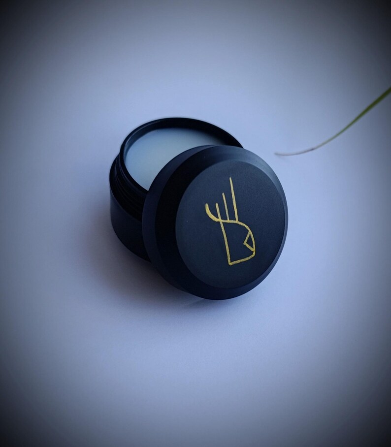 K&ouml;nnte beinhalten: Ein runder schwarzer Beh&auml;lter mit einem goldenen Design auf dem Deckel. Der Beh&auml;lter ist ge&ouml;ffnet und zeigt eine wei&szlig;e Creme im Inneren.