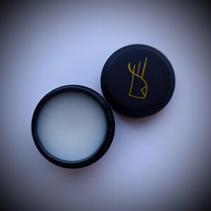 K&ouml;nnte beinhalten: Ein schwarzer Beh&auml;lter mit einer wei&szlig;en Creme darin. Der Deckel ist schwarz mit einem goldenen Logo eines stilisierten Hirschkopfes.