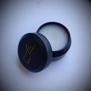 K&ouml;nnte beinhalten: Ein runder schwarzer Beh&auml;lter mit einem goldenen Logo eines stilisierten Fisches. Der Beh&auml;lter ist ge&ouml;ffnet und der Deckel liegt daneben. Der Beh&auml;lter ist mit einer wei&szlig;en Substanz gef&uuml;llt.