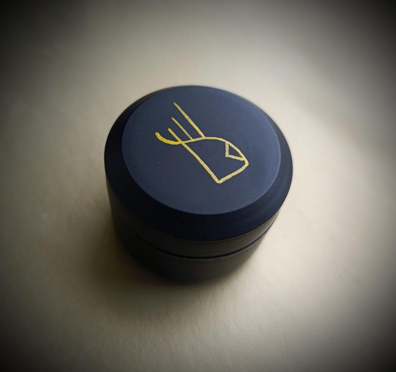 K&ouml;nnte beinhalten: Ein kleiner, runder, dunkelblauer Beh&auml;lter mit einem goldenen Design auf dem Deckel. Das Design ist ein stilisiertes Auge mit drei Linien, die daraus herausragen.