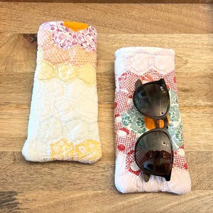 Puede incluir: Dos estuches rectangulares de tela acolchada con diseños de patchwork en tonos pastel de rosa, amarillo y blanco. Un estuche está vacío, mientras que el otro sostiene un par de gafas de sol con montura negra y lentes oscuras.