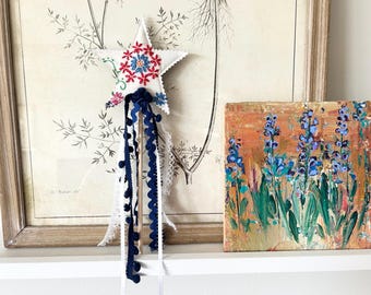May include: A white star-shaped wand with red, blue, and pink floral embroidery and navy blue and white ribbons. An abstract painting of blue flowers on an orange background is next to the wand. A framed botanical print is in the background.