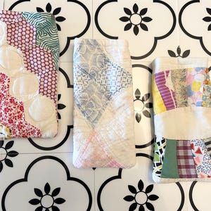 Puede incluir: Tres estuches rectangulares de tela acolchada para gafas u objetos pequeños. El estuche izquierdo presenta una mezcla de patrones florales y geométricos rojos, blancos y verdes. El estuche central tiene un diseño de rombos blancos y azules. El estuche derecho muestra un colorido patchwork de varios retazos de tela.