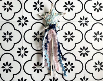 May include: A star-shaped wand with a light blue floral pattern, adorned with ribbons and lace in shades of pink, blue, and white. The wand is made of fabric and wood, perfect for imaginative play. The background is a white tile with black floral patterns.