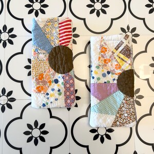 Puede incluir: Dos estuches rectangulares de tela acolchada con un diseño de patchwork de varios patrones florales, de rayas y geométricos en tonos azul, amarillo, rojo y blanco, cada uno centrado con un detalle de aplicación de tela marrón oscuro circular.