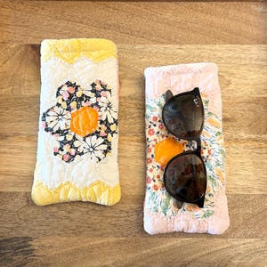 Puede incluir: Dos estuches rectangulares de tela acolchada para gafas. El estuche izquierdo presenta un fondo blanco con un gran estampado floral amarillo, negro y naranja. El derecho tiene un borde rosa pálido y un estampado floral con gafas de sol.