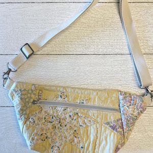 May include: A quilted fanny pack featuring a yellow star-patterned fabric with floral accents in shades of green, purple, and orange. The bag includes a horizontal silver zipper closure and an adjustable off-white woven strap with metal hardware.